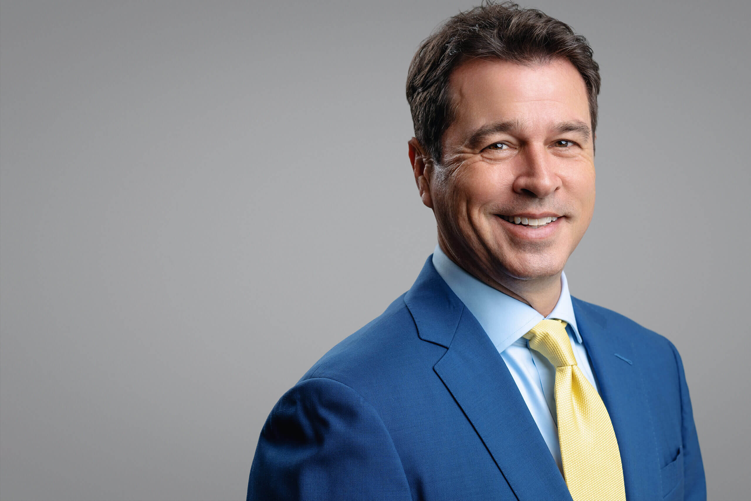 Smiling man in a blue suit and yellow tie against a gray background.