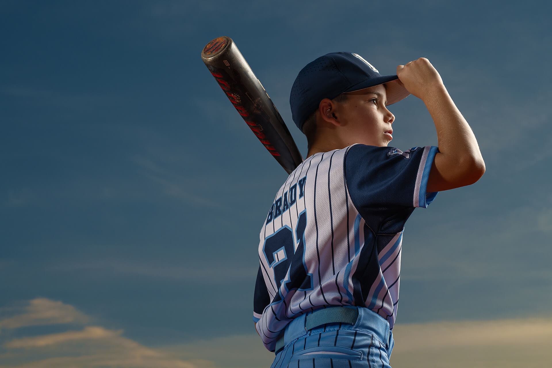 Young baseball player holding a bat against a cloudy sky.