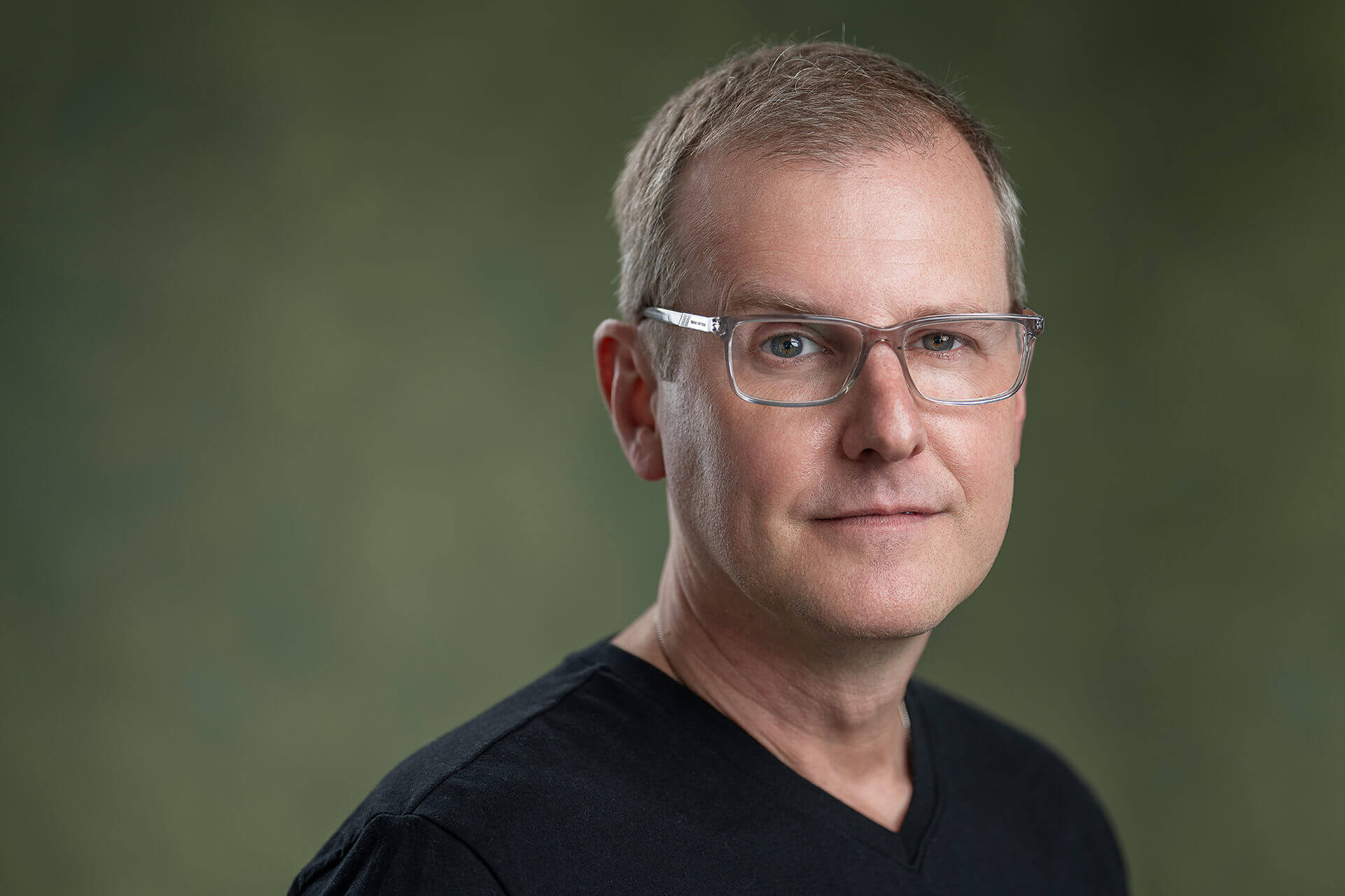 Man wearing glasses and a dark collared shirt against a green background.